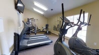 This single-floor fitness center offers a practical and well-equipped space for residents or members to maintain an active lifestyle. The room features warm beige walls and neutral carpeted flooring, creating a clean and functional environment. Overhead fluorescent lighting ensures the space is well-lit throughout the day, while a window fitted with horizontal blinds along one wall allows natural light to filter in.

The cardio section is prominently featured, housing Vision Fitness treadmills and an elliptical trainer positioned toward the front and side of the room. A stationary upright bike occupies the mid-section of the space, providing additional low-impact workout options. Toward the back wall, a multi-use cable and weight machine offers a range of strength training exercises, making this a versatile setup despite the room's modest footprint.

A large mirror panel lines the back wall, visually expanding the space and allowing users to monitor their form during workouts. A wall-mounted television is also present, adding convenience for entertainment during exercise sessions. Overall, the fitness center is straightforward and efficiently arranged.