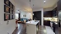 This thoughtfully designed single-floor residence offers a seamless open-concept layout that connects the kitchen, dining, and living areas into one cohesive living space. The kitchen is a standout feature, showcasing floor-to-ceiling dark espresso cabinetry, stainless steel appliances including a gas range, built-in microwave, and a French door refrigerator, all anchored by a large center island with a crisp white countertop, undermount sink, and seating for two. Elegant pendant lighting illuminates the island, while a track lighting system provides additional ambiance throughout. Rich, dark hardwood-style flooring flows throughout the main living areas, adding warmth and contrast to the light-toned walls. The living room is an inviting space, featuring a ceiling fan, natural light from a window with lush outdoor views, and room for comfortable furnishings. The bedroom continues the hardwood flooring and includes a ceiling fan, a window with natural light, and ample space for a full suite of furniture. The bathroom is clean and contemporary, offering a wide vanity with dark cabinetry and white countertop, a soaking tub with tile surround, and a separate glass-enclosed shower, all finished with crisp white tile flooring.