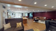 The West Lounge is a thoughtfully designed communal space that seamlessly blends comfort and functionality across a single open floor. Warm hardwood flooring runs throughout, tying together a series of distinct zones that cater to a variety of uses and group sizes.

At the heart of the space is a central kitchenette and bar area, featuring sleek grey countertops, white cabinetry, a built-in sink, and a long wooden communal table lined with industrial-style bar stools — ideal for casual meetings or social gatherings. A wall-mounted flat-screen TV anchors one end of this zone, complemented by decorative mirrors and tasteful accent pieces.

The lounge area is defined by its bold deep-crimson accent walls detailed with subtle horizontal banding, creating a warm and inviting atmosphere. A large L-shaped brown leather sectional sofa, a rustic wooden coffee table, and a geometric-patterned area rug furnish this cozy seating nook, complete with its own wall-mounted display screen.

A secondary seating zone features a darker, more intimate ambiance with charcoal walls, flexible seating including canvas chairs and a bold red accent bench, and a cylindrical side table. Natural light filters in through several windows, brightening the space. Framed artwork adorns the walls throughout, adding a refined, curated character to the overall environment.