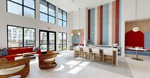 Located at 3280 Champion Ring Rd in Fort Myers, Florida, this single-floor community clubhouse offers an expansive and thoughtfully designed amenity space. Soaring double-height ceilings define the open-concept layout, creating an airy and grand atmosphere throughout. The main lounge area features plush, contemporary seating arrangements—including a large tufted white sectional and vibrant accent sofas in bold reds and teals—anchored by woven pendant light fixtures and a striking textured accent wall. Large floor-to-ceiling windows and black-framed glass doors flood the interiors with natural light while offering views of the surrounding community and pool area. A dedicated co-working zone provides individual workstations accented by teal feature walls and dramatic red pendant lighting, ideal for productivity. A relaxed reading nook with built-in shelving and soft lounge chairs offers a quieter retreat. The space also includes a game room complete with a pool table and a private golf simulator room for recreation. Bright tile flooring transitions seamlessly between zones, while patterned carpeting adds warmth to select areas. A casual dining nook with bar-height seating sits beneath expansive windows, completing this well-rounded, resort-style facility.