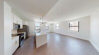 This contemporary single-floor residence offers a thoughtfully designed open-concept layout that seamlessly connects the kitchen and living areas. The kitchen is a standout feature, equipped with stainless steel appliances including a French door refrigerator, built-in microwave, and electric range. White shaker-style cabinetry with gold hardware extends to the ceiling, complemented by a generous white quartz island that doubles as a breakfast bar. Stylish globe pendant lights hang above, adding a modern touch to the space. The expansive living area is bathed in natural light streaming through multiple black-framed windows that frame urban skyline views, creating a bright and airy atmosphere throughout. Neutral-toned flooring flows through the main living spaces, while plush carpet adds comfort to the bedrooms. Each bedroom is well-proportioned, offering ample space and city views through large windows. The unit includes two bathrooms, both featuring classic black-and-white hexagonal mosaic tile flooring, white vanities with undermount sinks, and clean, minimalist finishes. One bathroom includes a walk-in glass-enclosed shower, while both offer bright, well-lit mirror arrangements. The overall design aesthetic is clean and contemporary, with a neutral palette that creates a versatile and welcoming environment.