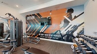 Located at 6490 Iron Horse Blvd in North Richland Hills, TX, this fitness center offers a well-equipped, single-floor exercise space designed to motivate and support a full range of workout routines. The room is defined by its bright, open feel, thanks to large black-framed floor-to-ceiling windows that flood the space with natural light and provide views of the surrounding outdoor pool area. Ceiling fans and recessed lighting ensure a comfortable environment during any workout.

A striking full-wall graphic mural serves as the centerpiece of the room, featuring bold orange, black, and white tones with the motivational message "LIMITLESS" and "We don't need to be perfect… we just need to get started." A wall-mounted flat-screen TV is positioned above the mural for entertainment during workouts. Floor-to-ceiling mirrors along one wall visually expand the space and support form-checking during exercise.

The fitness center is outfitted with a comprehensive selection of equipment, including treadmills, ellipticals, stationary and Echelon cycling bikes, a multi-station cable machine, a dumbbell rack with weight bench, and functional training accessories such as a heavy tire. The flooring features durable, textured carpet tile suited for high-traffic athletic use. Black-framed glass doors provide stylish transitions into and out of the space, completing its modern, purposeful aesthetic.