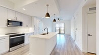 Located along Tamiami Trail East in Naples, Florida, this single-floor residence showcases a clean, contemporary design characterized by an all-white palette, light wood-style flooring, and matte black hardware accents throughout. The open-concept kitchen serves as the centerpiece of the living area, featuring a generously sized island with a quartz countertop, an undermount sink with a matte black faucet, and stainless steel appliances including a French door refrigerator, range, built-in microwave, and dishwasher. White shaker-style cabinetry extends to the ceiling, maximizing storage. Pendant lighting with dark metal shades hangs above the island, complementing the modern aesthetic. The kitchen flows seamlessly into a spacious living area illuminated by natural light from large black-framed windows and a sliding glass door that opens to a balcony. Ceiling fans are present throughout. The primary bathroom offers a long vanity with quartz countertops, a glass-enclosed shower, and direct access to a walk-in closet with built-in shelving. Additional bedrooms are bright and well-proportioned, each with ample natural light and closet access. The overall ambiance is clean, airy, and move-in ready.