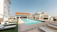 This rooftop amenity level offers a well-appointed outdoor retreat designed for relaxation and recreation. The centerpiece is a generously sized swimming pool with crystal-clear blue water, surrounded by elegant stone-tiled decking that provides ample space for lounging. A row of modern chaise lounges lines the pool's perimeter, offering comfortable seating under the open sky. A covered cabana structure with a distinctive terracotta-tiled roof anchors one end of the pool area, providing shaded space ideal for outdoor dining or social gatherings. The cabana appears to feature a built-in grill or outdoor kitchen, making it well-suited for entertaining. Pergola-style structures extend along the rooftop, adding architectural definition while framing panoramic views of the surrounding urban skyline. Lush artificial turf accents create a vibrant contrast against the neutral stone pavers, lending a resort-like ambiance to the space. The rooftop's elevated position affords expansive open-sky views and a sense of privacy above the city. The overall design blends functional amenities with refined architectural details, creating an inviting outdoor environment with a distinctly upscale character.
