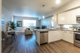 This well-designed single-floor residence offers a seamless open-concept layout that connects the kitchen, dining, and living areas into one cohesive, light-filled space. Rich dark hardwood-style flooring flows throughout the main living areas, creating a warm contrast against the cool grey walls and white trim.

The kitchen is a standout feature, outfitted with full stainless steel appliances including a refrigerator, electric range, microwave, and dishwasher. Sleek white flat-front cabinetry extends to the ceiling, maximizing storage, while under-cabinet lighting and a peninsula with pendant lighting add both function and style.

The open living and dining area is generously proportioned, anchored by large windows that invite natural light and views of surrounding greenery. A dedicated dining nook and spacious seating area provide flexible living arrangements.

The bedroom is a calm retreat, featuring plush carpet, a wide window with slatted blinds that allow for natural light, and ample wall space. The bathroom is finished with a neutral-toned countertop, an undermount sink, and a large mirror that enhances the sense of space.

Throughout, the design maintains a clean, contemporary aesthetic with consistent finishes and a bright, welcoming ambiance.