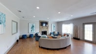 This single-story clubhouse offers a thoughtfully designed communal lounge space that serves as a central gathering point for residents. The main living area is anchored by a striking floor-to-ceiling stacked stone fireplace, above which a wall-mounted flat-screen television is positioned for easy viewing from multiple seating arrangements. Rich hardwood flooring extends throughout the space, adding warmth and a polished finish to the interior.

The lounge is furnished with a large, curved sectional sofa in a neutral tone, complemented by coordinating blue accent chairs and decorative ottomans that introduce pops of color. Throw pillows in teal, orange, and gray tie the color palette together cohesively. Two large abstract artworks in shades of blue flank the fireplace, contributing to the space's contemporary aesthetic.

Recessed lighting is evenly distributed across the white ceiling, providing ample illumination throughout. Crown molding adds a refined architectural detail. A window dressed with sheer curtains allows natural light to filter into the room, enhancing the open and inviting ambiance of this well-appointed community clubhouse.
