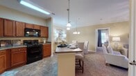 This single-floor residence offers a thoughtfully designed layout that flows seamlessly from room to room. The entryway, accented by decorative artwork and a console table with a mirror, leads into the main living areas. The kitchen is equipped with rich cherry wood cabinetry, black appliances including a range, microwave, and refrigerator, and granite-style countertops. A central island with pendant lighting and bar seating serves as a functional hub, connecting directly to the dining area, which features an additional chandelier and seating for several guests. The living room is carpeted and benefits from natural light through windows and a glass door leading to an outdoor area, complemented by a ceiling fan with light fixture and crown molding details. Two bedrooms feature plush carpeting, natural light from windows with blinds and draping curtains, and warm golden-toned walls. The bathroom includes a shower-tub combination, wood-toned vanity cabinetry, and tile flooring. Throughout, consistent warm beige tones, crown molding, and tile-to-carpet transitions create a cohesive and comfortable atmosphere.