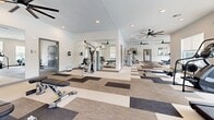 Located at 18905 E 59th Ave in Denver, CO, this modern fitness center spans a generous single floor and is divided into two dedicated exercise rooms, offering a comprehensive workout environment for users of all fitness levels. The space features a clean, contemporary design with neutral-toned walls, patterned carpet tile flooring in varying shades of beige, tan, and charcoal, and recessed lighting complemented by large, modern ceiling fans that ensure comfortable air circulation throughout. Natural light streams in through multiple windows fitted with roller shades, with views overlooking an outdoor pool area. Full-length mirrors are strategically positioned to enhance the sense of openness and support proper form during training. The main exercise room is outfitted with a diverse selection of strength and cardio equipment, including treadmills, cable machines, weight benches, a dumbbell rack, a rowing machine, and an elliptical trainer. Wall-mounted Samsung flat-screen televisions add an entertainment element to the workout experience. A dedicated functional training area houses a multi-purpose rack stocked with resistance bands, medicine balls, kettlebells, and TRX suspension trainers. The second exercise room provides additional cardio equipment such as stationary bikes, an elliptical, and open floor space ideal for stretching or plyometric training, including a plyo box.