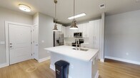 This single-floor residence showcases a thoughtfully designed layout with modern finishes throughout. The heart of the home is an open-concept kitchen, where crisp white cabinetry extends floor to ceiling, providing generous storage. A classic white subway tile backsplash complements the stainless steel appliance suite, which includes a refrigerator with ice and water dispenser, an electric range, and an over-the-range microwave. A large central island with a bright white countertop and an integrated stainless steel sink serves as both a functional workspace and a natural gathering point. Overhead, brushed brass pendant lights add warmth and a contemporary accent to the space. Light wood-tone flooring runs throughout the main living and kitchen areas, contributing to an airy, cohesive aesthetic. The bedroom is a calm, well-lit retreat featuring plush carpeting and multiple windows that frame lush outdoor views, filling the room with natural light. The bathroom is finished with clean white subway tile surrounding a full soaking tub and shower combination, brushed nickel fixtures, and wood-look flooring that ties the space together with understated elegance. Overall, the residence presents a bright, modern atmosphere with consistent attention to detail in its finishes and fixtures.