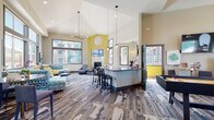 Located at 483 Scott Blvd in Castle Rock, Colorado, this single-story community clubhouse delivers a lively and welcoming atmosphere designed for both relaxation and recreation. The space is defined by dramatically soaring vaulted ceilings that amplify its open, airy feel throughout.

The main lounge area is generously proportioned, featuring multiple distinct seating arrangements with modern, colorful furniture including yellow accent chairs, blue leather seating, and patterned sofas set atop bold, multi-toned area carpeting. A tiled fireplace anchors one wall, topped by a mounted flat-screen television, while large floor-to-ceiling windows flood the space with abundant natural light and offer views of the surrounding community courtyard.

An interior bar-height counter with dark countertops and industrial-style pendant lighting provides a central gathering point. Adjacent to the lounge, a recreation area features a shuffleboard table, arcade games, and additional entertainment screens, creating an energetic social hub.

A dedicated business center nook offers a height-adjustable desk, computer workstation, and ample seating for residents needing a quiet workspace. The full kitchen is equipped with wood-grain cabinetry, stainless steel appliances including a dishwasher and wine refrigerator, a tiled backsplash, and generous counter space. Bold yellow accent walls and curated artwork throughout contribute to the space's contemporary, community-focused character.