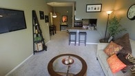 This single-floor residence showcases an open-concept layout that connects the living room, dining area, and kitchen in one cohesive space. The living room is anchored by a round wooden coffee table with a glass inset, complemented by plush carpeting that extends throughout the main areas. Sage green walls lend a calm, neutral tone to the interior. A decorative ladder shelf adds a stylish organizational element along one wall, while framed artwork contributes to the curated aesthetic.

The kitchen is partially separated by a raised breakfast bar with a dark stone countertop, accommodated by two dark bar stools. The kitchen features dark appliances and ample counter space, with a window that draws in natural light. A pass-through opening between the kitchen and living area enhances the open feel and allows easy interaction across spaces.

Beyond the kitchen, the dining area accommodates a dark wood table and chairs beneath a classic pendant light, creating a defined yet connected eating space. The overall ambiance is warm, inviting, and functional, with consistent flooring and wall colors tying each zone together cohesively.
