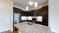 Located at 1005 West 5th Avenue in Columbus, Ohio, this single-floor residence offers a thoughtfully designed living space that blends modern aesthetics with everyday functionality. The kitchen serves as a standout feature, showcasing rich dark-toned cabinetry that extends to the ceiling for generous storage capacity. Gleaming granite countertops with a speckled finish provide ample workspace and continue onto a central island equipped with a built-in stainless steel sink — an ideal setup for both cooking and casual dining. A full suite of stainless steel appliances, including a refrigerator, range, and microwave, reinforces the kitchen's contemporary character. A classic white subway tile backsplash adds a clean contrast against the dark cabinetry, while pendant lighting suspended above the island delivers focused illumination and an element of modern style. The overall design is cohesive and polished, with a neutral color palette that creates a bright, welcoming ambiance throughout. This single-level space is well-suited for those seeking a refined, move-in ready home in the heart of Columbus's vibrant West Side neighborhood.