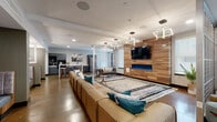 This single-floor amenity space draws inspiration from Nashville's vibrant culture, incorporating locally themed black-and-white artwork throughout. The layout flows seamlessly between distinct zones, each designed for a specific type of social or recreational activity.

The main lounge area anchors the space with a large sectional sofa, a striking wood-paneled feature wall, a wall-mounted TV, and a sleek electric fireplace glowing in blue. Geometric pendant lighting overhead adds a modern design touch. Adjacent to this is an open kitchenette outfitted with gray cabinetry, stainless steel appliances, a white countertop island, and bar seating.

A dedicated dining area features upholstered booth seating set against dramatic large-scale abstract murals, complemented by sputnik-style pendant fixtures. Nearby, a casual seating nook furnished with plush navy and white chairs is adorned with Nashville-themed photography, creating an inviting gathering spot.

The game room offers a full-size pool table, a wood-paneled TV wall with shelving, and a water station set against a bold navy accent wall. Additional areas include a versatile open room with a built-in work surface, a living wall installation, a barn-style sliding door, and access to an outdoor terrace. Polished concrete flooring runs throughout, tying the entire space together with an industrial-modern aesthetic.