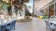 This inviting outdoor courtyard is nestled between two multi-story historic brick buildings that lend the space a rich, industrial character. The exposed brick facades, accented by large factory-style windows and climbing vines, frame the courtyard with a timeless architectural presence that speaks to the building's storied past.

A central stone-edged fountain serves as the focal point of the courtyard, surrounded by a broad concrete pathway that flows the length of the space. Mature magnolia and deciduous trees provide generous shade and natural beauty throughout, while carefully maintained landscaping — including ornamental shrubs, gravel beds, and ground cover plantings — softens the hardscape and adds color and texture.

The courtyard is thoughtfully furnished for outdoor enjoyment, featuring multiple seating zones with vibrant yellow and teal Adirondack chairs, traditional wooden benches, and dining-style patio furniture. A grill station adds functional outdoor amenity, making the space well-suited for social gatherings.

The entrance is framed by an elegant iron gate, a covered entry structure, and additional greenery that creates a welcoming transition from the street. Throughout, the combination of historic architecture, mature trees, and colorful modern furnishings creates a warm, communal ambiance that balances urban energy with a relaxed, park-like atmosphere.