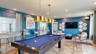 Located in Naples, FL, this single-story community clubhouse at Sumter Grove Circle offers a thoughtfully designed gathering space with a bold, contemporary aesthetic. The grand entrance lobby immediately impresses with soaring vaulted ceilings adorned with dark wood beam accents and an eye-catching geometric chandelier. Rich crimson armchairs arranged around a striking gold-and-black coffee table create an inviting seating area, while a vivid teal accent wall with gold patterned wallcovering and an ornate mirror adds dramatic flair. Abundant natural light pours in through large windows, enhancing the open, airy atmosphere. Adjacent to the lobby, the game and lounge area showcases deep teal walls, intricate parquet-style wood flooring, and a regulation billiards table illuminated by oversized gold pendant lights. High-top bar tables with gold-accented chairs provide additional social gathering spots, complemented by curated geometric artwork. A dedicated lounge nook features a wall-mounted flat-screen TV surrounded by hexagonal decorative frames, flanked by stylish accent chairs over a richly patterned area rug. Throughout the space, patterned drapery, lush tropical plants, and carefully selected art pieces contribute to a cohesive, resort-inspired ambiance that reflects the spirit of Southwest Florida living.