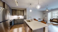This well-appointed single-floor residence showcases a thoughtfully designed open-concept layout that seamlessly connects the kitchen, dining, and living areas. The kitchen is a standout feature, outfitted with rich dark-toned shaker-style cabinetry, stainless steel appliances including a range, microwave, and refrigerator, and a crisp white subway tile backsplash. A large center island with a light quartz countertop, undermount sink, and seating for two serves as the heart of the space, illuminated by a pair of stylish globe pendant lights. Wood-look flooring flows throughout the main living areas, complementing the warm, contemporary aesthetic. The living room offers an inviting atmosphere with an arched architectural detail, a ceiling fan, and windows that bring in natural light and neighborhood views. The bedroom is carpeted for comfort and features a generous walk-in closet with built-in shelving and a ceiling fan. The bathroom is clean and bright, with marble-look tile flooring, a tub-shower combination, and white fixtures that create a crisp, minimalist finish. Built-in shelving in the living area and track lighting in the kitchen add both function and character to this modern, move-in-ready space.