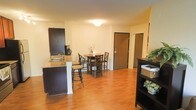 This single-floor space is designed with an open-concept layout that seamlessly connects the kitchen, dining, and living areas, creating a sense of flow and spaciousness throughout. The warm-toned hardwood flooring extends across the entire main living area, lending a clean and inviting aesthetic.

The kitchen is outfitted with dark cabinetry that provides a striking contrast against the light walls. A peninsula with a granite-style countertop and bar seating serves as both a functional workspace and a casual dining spot, making it an ideal area for everyday meals or entertaining. The kitchen is equipped with standard appliances, including a coffee maker and kettle, and features a stainless steel sink.

Adjacent to the kitchen, the dedicated dining area accommodates a round table with cross-back chairs, creating a cozy and defined space for shared meals. The entry door is conveniently located nearby, making the layout practical and easy to navigate.

The light-colored walls and flush ceiling lighting keep the interior bright and airy, while a dark bookshelf unit near the living area adds storage and a touch of contrast. Overall, the space offers a well-balanced and functional design with a warm, comfortable ambiance suited for everyday living.