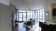 This stylish single-floor residence makes an immediate impression with its expansive floor-to-ceiling window walls that flood the interior with abundant natural light and frame sweeping panoramic views of the surrounding cityscape and skyline. The open-concept living area is anchored by rich, dark hardwood flooring that contrasts beautifully with the bright, white ceiling and bold accent walls painted in a deep navy blue. Large black-framed glass panels and French doors provide direct access to the outdoors, further blurring the line between interior and exterior living. A sleek ceiling fan with a brushed metal finish adds both style and functionality to the space. The living room is thoughtfully arranged with a curated mix of contemporary and transitional furnishings, complemented by patterned drapery and a decorative area rug that add texture and visual interest. Accent furniture and decor items are tastefully integrated throughout. The overall ambiance is one of modern urban sophistication — bright, open, and elegantly appointed — with the dramatic window walls serving as the defining architectural feature that draws the eye outward toward expansive city views.
