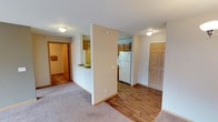 This single-floor residential unit offers a practical and well-organized layout designed for comfortable everyday living. The open floor plan connects the main living area to the kitchen, creating a cohesive and accessible flow throughout the space. The living area is finished with soft wall-to-wall carpeting, while the kitchen and entryway transition to warm hardwood-style flooring, adding visual contrast and easy maintenance underfoot. The kitchen is equipped with wood-toned cabinetry, a white refrigerator, and a countertop with a sink, providing a functional workspace for meal preparation. A half-wall partition subtly separates the kitchen from the living space while maintaining an open, airy feel. Flush-mount ceiling light fixtures provide consistent illumination throughout the unit. A wood-panel door near the kitchen area serves as the main entry point, offering convenient access to and from the unit. A bathroom is accessible from the main living area, featuring tiled flooring and standard fixtures. The neutral color palette of soft whites and warm beige tones throughout the unit creates a clean, inviting atmosphere that is easy to personalize. Overall, the space is thoughtfully laid out to maximize functionality within a compact single-level footprint.