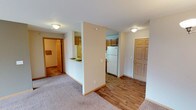 This single-floor residential unit offers a practical and well-organized layout designed for comfortable everyday living. The open floor plan connects the main living area to the kitchen, creating a cohesive and accessible flow throughout the space. The living area is finished with soft wall-to-wall carpeting, while the kitchen and entryway transition to warm hardwood-style flooring, adding visual contrast and easy maintenance underfoot. The kitchen is equipped with wood-toned cabinetry, a white refrigerator, and a countertop with a sink, providing a functional workspace for meal preparation. A half-wall partition subtly separates the kitchen from the living space while maintaining an open, airy feel. Flush-mount ceiling light fixtures provide consistent illumination throughout the unit. A wood-panel door near the kitchen area serves as the main entry point, offering convenient access to and from the unit. A bathroom is accessible from the main living area, featuring tiled flooring and standard fixtures. The neutral color palette of soft whites and warm beige tones throughout the unit creates a clean, inviting atmosphere that is easy to personalize. Overall, the space is thoughtfully laid out to maximize functionality within a compact single-level footprint.