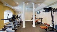 This fitness center offers a thoughtfully designed workout environment that balances functionality with classic architectural charm. The space is defined by its prominent white decorative columns, which rise from floor to ceiling and lend a refined aesthetic to an otherwise utilitarian setting. The walls are finished in a warm neutral tone, complemented by dark, durable flooring suited for high-traffic exercise use. Recessed lighting throughout the ceiling ensures the space is well-lit, while a ceiling fan provides additional air circulation during workouts. The cardio zone lines one side of the room with a row of treadmills and elliptical machines, offering ample options for aerobic training. On the opposite side, a selection of strength and resistance training equipment — including a cable machine and an adjustable weight bench — provides users with a comprehensive range of exercise options. A wall-mounted television is positioned to keep users entertained during their sessions. An emergency exit sign and clearly marked door contribute to the safety and practicality of the layout. Overall, the fitness center delivers a clean, organized, and inviting atmosphere suitable for a full range of workout routines.
