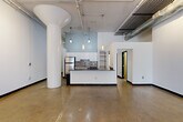 Located at 820-828 Ralph McGill Blvd NE in Atlanta's Old Fourth Ward neighborhood, this single-floor loft-style space offers a generous and versatile layout with a distinctive industrial aesthetic. Polished and raw concrete floors run throughout, complemented by crisp white walls and bold black trim accents on doors and baseboards. The open main living area is anchored by striking cylindrical structural columns and features soaring ceilings with exposed ductwork, pipes, and track lighting, creating a dynamic urban atmosphere. A built-in kitchen includes white cabinetry, a dark-toned island with pendant lighting above, a mosaic tile backsplash, and stainless steel appliances. A private bedroom area is served by large steel-framed multi-pane windows that invite abundant natural light and views of surrounding greenery, with a ceiling fan adding functional comfort. A bathroom includes a modern vanity with an undermount sink and stone-look countertop, alongside natural wood-toned doors with black frames. A long interior corridor connects the spaces efficiently, while an additional room with distressed concrete floors offers further flexibility. The overall ambiance is open, light-filled, and raw — ideal for creative or residential use.