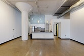 Located at 820-828 Ralph McGill Blvd NE in Atlanta's Old Fourth Ward neighborhood, this single-floor loft-style space offers a generous and versatile layout with a distinctive industrial aesthetic. Polished and raw concrete floors run throughout, complemented by crisp white walls and bold black trim accents on doors and baseboards. The open main living area is anchored by striking cylindrical structural columns and features soaring ceilings with exposed ductwork, pipes, and track lighting, creating a dynamic urban atmosphere. A built-in kitchen includes white cabinetry, a dark-toned island with pendant lighting above, a mosaic tile backsplash, and stainless steel appliances. A private bedroom area is served by large steel-framed multi-pane windows that invite abundant natural light and views of surrounding greenery, with a ceiling fan adding functional comfort. A bathroom includes a modern vanity with an undermount sink and stone-look countertop, alongside natural wood-toned doors with black frames. A long interior corridor connects the spaces efficiently, while an additional room with distressed concrete floors offers further flexibility. The overall ambiance is open, light-filled, and raw — ideal for creative or residential use.