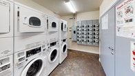 This communal laundry room is a well-organized, single-floor facility designed to serve multiple users efficiently. The room houses several stacked washer and dryer units — commercial-grade front-loading machines arranged in rows along the walls — providing ample capacity for shared use. The machines are clearly labeled and feature digital controls, with posted instructional guides for washing and drying displayed on the wall for easy reference.

The space features durable, dark concrete-style flooring that is both practical and easy to maintain. Neutral-toned walls keep the environment clean and functional in appearance. Overhead fluorescent lighting ensures the room is well-lit throughout, while a window along one wall allows for natural light and ventilation.

A utility panel with multiple meters is mounted on one wall, reflecting the building's shared infrastructure. A waste bin and basic supplies are accessible within the room for user convenience. The room is accessible through a standard door, making entry and exit straightforward.

Overall, this laundry room is designed with practicality and efficiency in mind, offering a clean, functional environment with all the essential amenities needed for a well-equipped communal laundry facility.