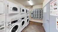 This communal laundry room is a well-organized, single-floor facility designed to serve multiple users efficiently. The room houses several stacked washer and dryer units — commercial-grade front-loading machines arranged in rows along the walls — providing ample capacity for shared use. The machines are clearly labeled and feature digital controls, with posted instructional guides for washing and drying displayed on the wall for easy reference.

The space features durable, dark concrete-style flooring that is both practical and easy to maintain. Neutral-toned walls keep the environment clean and functional in appearance. Overhead fluorescent lighting ensures the room is well-lit throughout, while a window along one wall allows for natural light and ventilation.

A utility panel with multiple meters is mounted on one wall, reflecting the building's shared infrastructure. A waste bin and basic supplies are accessible within the room for user convenience. The room is accessible through a standard door, making entry and exit straightforward.

Overall, this laundry room is designed with practicality and efficiency in mind, offering a clean, functional environment with all the essential amenities needed for a well-equipped communal laundry facility.