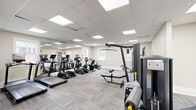 Located at 750 Chateau Ln in Riverdale, Georgia, this fitness center offers a well-appointed exercise space designed to support a comprehensive workout experience. The facility is housed on a single floor and features a clean, modern aesthetic with neutral-toned walls, white-trimmed doors, and patterned carpet tile flooring throughout.

The main exercise area is thoughtfully organized into distinct zones. The cardio section is equipped with multiple TRUE brand treadmills and an elliptical machine, positioned along a mirrored wall that enhances the sense of openness. Adjacent to the cardio area, a dedicated strength training zone houses a multi-station cable machine and a leg extension/curl unit, providing a variety of resistance training options. A selection of stability and medicine balls rounds out the functional fitness area.

Natural light filters in through several windows, complementing the bright, recessed panel lighting in the drop ceiling above. A glass-panel sliding door provides a transparent boundary between the entry vestibule and the main workout floor, contributing to an airy, open feel. A water station is conveniently accessible within the space. The overall layout is spacious and uncluttered, creating an inviting and functional environment well-suited for a range of fitness routines.