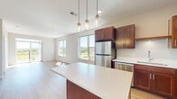 Located at 1875 Paulson Rd in River Falls, Wisconsin, this thoughtfully designed one-bedroom, one-bathroom unit offers a comfortable and modern living experience across a single floor. The open-concept kitchen and living area serves as the heart of the unit, where rich cherry-toned cabinetry, crisp white quartz countertops, and a full suite of stainless steel appliances — including a refrigerator, range, microwave, and dishwasher — create a stylish and functional culinary space. A center island with pendant lighting provides additional prep space and casual seating, while light hardwood-style flooring flows throughout the main living areas, lending warmth and continuity to the design. Large windows in the living room flood the space with natural light and offer open views of the surrounding landscape, enhancing the airy, spacious feel of the unit. The bedroom is carpeted for comfort, features its own window with outdoor views, and includes a dedicated closet. The full bathroom is finished with matching cherry cabinetry, white countertops, a tub-shower combination, and hardwood-style flooring that ties seamlessly to the rest of the unit. A hallway connects the rooms efficiently, completing this clean and cohesive layout.