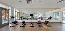 This contemporary fitness studio is thoughtfully designed to support a range of workout activities across a single, well-appointed floor. The space is divided into two primary zones: a dedicated indoor cycling room and an open multipurpose studio area.

The cycling room is outfitted with a full row of professional-grade stationary bikes arranged in front of a large wall-to-wall mirror, which amplifies the sense of space and allows participants to monitor their form. A mounted flat-screen display serves as a focal point for instructor-led or on-demand fitness programming. The warm hardwood-style flooring and white walls create a clean, energizing atmosphere throughout.

The adjacent open studio area provides ample floor space for group fitness classes, yoga, stretching, or personal training sessions. A "Fitness On Demand" display system is integrated into the mirrored wall, offering flexible, self-guided workout options. Floor-to-ceiling glass doors and expansive windows line one side of the studio, flooding the room with natural light and offering views of the outdoor surroundings, creating an open and inviting environment.

Both areas share consistent design elements — hardwood-style flooring, recessed and flush-mount lighting, and a bright, neutral color palette — resulting in a cohesive, professional fitness environment suited for a variety of training needs.