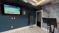 This well-appointed Yoga/Spin Studio is designed to accommodate a wide range of fitness activities within a single, thoughtfully organized space. The studio's interior is defined by bold deep-blue walls accented by a striking textured feature wall with a blue, gray, and gold marbled pattern, lending the space a dynamic and energizing aesthetic. Light-toned striped vinyl flooring runs throughout, providing a clean, durable surface ideal for high-intensity workouts.

The space is equipped with spin bikes positioned in front of the decorative accent wall, alongside an array of fitness accessories including stability balls, Smart Core training boards, resistance equipment, and a vibration platform. A wall of floor-to-ceiling mirrors on one side expands the sense of space and supports form monitoring during exercise. A large wall-mounted flat-screen television ensures participants can stay entertained or follow guided workouts during sessions.

Exposed industrial-style ductwork runs overhead, complemented by linear LED lighting and warm recessed cove lighting that creates an inviting atmosphere. A distinctive exposed brick column adds a textural contrast to the modern finishes. Glass-panel doors offer a visual connection to adjacent areas, while a potted fiddle-leaf fig tree adds a natural, refreshing touch. The studio is well-ventilated and designed to support a full range of group or individual fitness programming.