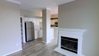 This single-floor space offers an open, connected layout that integrates the living area and kitchen into one cohesive environment. The living area is anchored by a white-trimmed fireplace with gold-accented glass doors and classic tile surround, serving as a standout focal point. Wood-style plank flooring runs throughout, adding warmth and visual continuity across the space.

The kitchen is well-appointed with white cabinetry, granite-look countertops, and stainless steel appliances, including a full-sized refrigerator and microwave. A peninsula counter with bar-height seating potential creates a natural transition between the kitchen and the main living area, supporting both functionality and an open feel. A structural column between the two zones adds architectural character while maintaining the open sightlines.

Neutral gray walls and white trim work together to create a clean, modern aesthetic throughout. Overhead vents and ample ceiling height contribute to an airy atmosphere. The overall design is practical and cohesive, making the space feel welcoming and move-in ready.