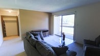 This single-floor space centers around a welcoming living room that sets a comfortable and functional tone throughout. The living area is highlighted by a large sliding glass door that floods the room with natural light and opens directly onto a private balcony, offering pleasant outdoor views of surrounding greenery. The carpeted floor adds warmth underfoot, while the textured ceiling and neutral-toned walls create a calm, understated atmosphere. The living room comfortably accommodates a plush sectional sofa paired with a glass-top coffee table and a classic wooden media console, demonstrating generous space for relaxed everyday living and entertaining. The balcony, enclosed by a metal railing, extends the living experience outdoors and provides a serene retreat with tree-lined views. The overall ambiance of the space is practical yet inviting, with an emphasis on natural light and ease of flow. The single-floor layout ensures seamless accessibility throughout, making it a straightforward and livable environment well-suited for day-to-day comfort.