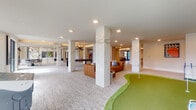 Located at 5201 Congress Ave in Boca Raton, Florida, this contemporary amenity space delivers an impressive blend of refined design and recreational offerings across a single, well-appointed floor. The grand entry lobby sets an elegant tone with light hardwood flooring, soaring floor-to-ceiling glass walls, a floating staircase with glass railings and warm wood treads, and a sculptural pendant light fixture. Marble-clad columns appear throughout, anchoring the open layout while adding a luxurious architectural detail. The centerpiece of the main gathering area is a full indoor putting green, surrounded by plush carpet, comfortable lounge seating in warm leather tones, and a golf simulator — a standout feature for sports enthusiasts. Adjacent areas include a game room with a billiards table, a dedicated poker/card table room bathed in natural light from expansive windows overlooking lush tropical greenery, and relaxed lounge nooks furnished with stylish modern seating. A wet bar and kitchenette area with bar seating provides a functional social hub. Wide-format tropical and contemporary artwork adorns the corridors and walls throughout, reinforcing the South Florida aesthetic. Large windows and glass doors connect interior spaces to outdoor views, filling the amenity floor with abundant natural light and an airy, resort-inspired ambiance.