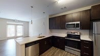 This single-floor, one-bedroom, one-bathroom unit in Memphis, TN's Cole neighborhood offers a well-designed, open layout with consistent hardwood-style flooring flowing throughout the main living areas. The kitchen is a standout feature, equipped with dark espresso cabinetry, light quartz-style countertops, and a full suite of stainless steel appliances, including a refrigerator, electric range, built-in microwave, and dishwasher. Stylish pendant lighting hangs above the peninsula, which provides a natural division between the kitchen and the open living area. The living space is generously sized and bright, featuring neutral walls and direct access to a private balcony through French doors, allowing natural light to fill the room. The bedroom offers plush carpeting, a large window with exterior views, and a panel-style door. The bathroom is cleanly finished with wood-style flooring, a full soaking tub, and tiled shower surround in a subtle gray palette. Overall, the unit presents a modern, cohesive aesthetic with neutral tones throughout.