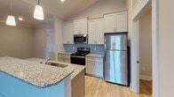 Located at 6030 Neal Trail Dr in Walkertown, North Carolina, this single-floor home offers a well-designed layout with a blend of modern finishes and functional living spaces. The open-concept kitchen is a standout feature, showcasing white shaker-style cabinetry extending to the ceiling, a light blue tile backsplash, stainless steel appliances including a refrigerator, range, and over-the-range microwave, and a granite-topped peninsula with an undermount sink — all set atop durable hardwood-style flooring with vaulted ceiling accents that add a sense of height and openness. The living area flows naturally from the kitchen, featuring plush neutral carpeting and a clean, airy aesthetic with warm beige tones throughout. Multiple bedrooms offer comfortable proportions, each with soft carpeting, ceiling fans with light fixtures, and windows fitted with white blinds that allow natural light. Two bathrooms are finished with granite countertops, gray shaker-style vanity cabinetry, large framed mirrors, and tile flooring — one features a tub/shower combo and the other includes a walk-in shower area. A dedicated laundry space provides washer and dryer hookups with built-in wire shelving above. The overall ambiance is bright, clean, and move-in ready, reflecting a thoughtfully updated interior throughout.