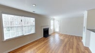 This single-floor space showcases a welcoming living area defined by warm, honey-toned hardwood flooring that reflects natural light throughout the room. A classic fireplace with a clean white surround serves as the room's focal point, adding both visual interest and functional warmth to the space. Crown molding runs along the ceiling perimeter, lending a refined architectural detail that elevates the overall aesthetic.

The room benefits from generous natural light, courtesy of multiple windows fitted with white horizontal blinds, allowing for adjustable brightness and privacy. The neutral, warm beige wall tones complement the flooring and create a versatile backdrop suitable for a variety of interior styles. Recessed lighting in the ceiling provides additional illumination, ensuring the space remains bright regardless of the time of day.

A door adjacent to one of the windows offers direct access to the exterior, enhancing both functionality and the sense of openness. The clean, simple lines throughout the space, combined with white baseboards and trim, contribute to a tidy, cohesive look that makes this living area feel inviting and move-in ready.
