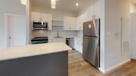 Located at 1 Royal Dr. in Somersworth, New Hampshire, this thoughtfully designed single-floor residence offers a clean, modern aesthetic with neutral tones and quality finishes throughout. The contemporary kitchen is a standout feature, equipped with crisp white cabinetry, open shelving, light quartz countertops, and a full suite of stainless steel appliances — including a refrigerator, over-the-range microwave, and dishwasher. A bold teal accent on the range adds a distinctive touch, while a central island with a contrasting gray base provides additional prep and dining space. Light wood-look flooring runs throughout the kitchen and connecting areas, contributing to a bright, cohesive feel. A carpeted bedroom offers a comfortable retreat, with a window overlooking a private balcony and surrounding greenery, allowing natural light to filter in. A spacious walk-in closet features multi-tier wire shelving and additional hanging space, providing ample storage. The bathroom is finished with clean white walls, light flooring, and chrome hardware. A well-lit hallway connects the living areas seamlessly, with white panel doors and recessed lighting maintaining the home's modern, polished character throughout.