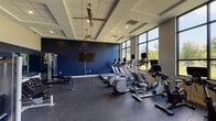 This fitness center is thoughtfully designed to support a comprehensive workout experience within a single, open-plan space. The room features elevated ceilings with a coffered design and ceiling fans, contributing to a spacious, airy atmosphere. Bold navy blue accent walls contrast with neutral tones throughout, giving the space a modern, energetic aesthetic. Durable rubber flooring covers the entire area, providing both safety and functionality underfoot.

Along the windowed wall, floor-to-ceiling large-pane windows flood the space with abundant natural light while offering views of the surrounding greenery outdoors. This wall accommodates a full lineup of cardio equipment, including treadmills, elliptical machines, stationary bikes, and a cycling bike, each equipped with individual monitors for an engaging workout experience.

The strength-training area features a tiered dumbbell rack with an extensive range of free weights, multiple adjustable workout benches, a cable machine, and additional selectorized strength equipment. A large wall mirror enhances the sense of depth and supports proper form during exercises. Colorful resistance bands, medicine balls, and foam rollers are also available, offering versatility for functional training. A dedicated entry area with a glass door provides direct outdoor access, completing this well-rounded facility.