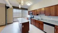This single-floor residential unit offers a well-organized layout that balances open living spaces with functional private rooms. The kitchen serves as a central highlight, featuring rich dark wood cabinetry, granite countertops, a large center island, and a full suite of stainless steel appliances including a refrigerator, range, microwave, and dishwasher. Light-toned flooring and large windows with horizontal blinds allow ample natural light to fill the space.

The living area is distinguished by warm hardwood flooring, a bold teal accent wall, track lighting along the ceiling, and expansive windows that further enhance the sense of openness. Bedrooms are carpeted in a neutral tone and equipped with ceiling fans fitted with lighting. Closet space is provided in the bedrooms, with one room featuring a built-in closet with bifold doors. A fire alarm strobe is also present, indicating compliance with safety standards.

Two bathrooms serve the unit: one includes a walk-in shower with grab bars and a dark wood vanity with a granite countertop, while the other offers a full tub-shower combination, a matching vanity, and a lighted mirror. Both bathrooms feature tile flooring and neutral wall tones. Overall, the unit presents a clean, modern aesthetic with consistent finishes throughout.