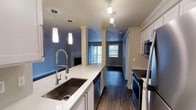 This well-appointed 3-bedroom, 2-bathroom residence offers a thoughtfully designed single-level layout that maximizes both comfort and functionality. The open-concept kitchen serves as the heart of the home, showcasing crisp white cabinetry, gleaming quartz countertops, and a large stainless steel undermount sink equipped with a professional-style coil faucet. Stainless steel appliances, including a range and dishwasher, complement the modern aesthetic, while track lighting and elegant pendant lights illuminate the space with a warm, inviting glow. Decorative columns with crown molding detail define the transition between the kitchen and the adjoining living area, adding a touch of architectural character. Rich, dark hardwood-style flooring runs throughout, providing a striking contrast to the light-colored cabinetry and walls painted in a cool, contemporary blue-gray tone. Crown molding accents the ceilings, further elevating the overall finish of the space. The living area benefits from natural light streaming in through windows that offer views of the outdoors. The combination of stylish finishes, functional layout, and attention to detail makes this residence a comfortable and visually appealing home.