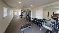 This single-floor fitness center is thoughtfully designed to support a comprehensive workout experience. The space is organized into distinct functional zones, each catering to different aspects of physical training. The cardio area features a row of treadmills and elliptical machines lined along a wall of floor-to-ceiling mirrors, which amplify the sense of space and allow users to monitor their form. Large windows and sliding glass doors along one side of the facility flood the space with natural light and provide views of the surrounding outdoor greenery. The strength training zone is well-appointed with a multi-station cable machine, a selection of weight benches, and a tiered dumbbell rack stocked with a wide range of free weights, all positioned beneath recessed ceiling lighting. A separate flexibility and functional fitness area offers ample open floor space and is stocked with stability balls in multiple sizes, resistance bands, and rolled exercise mats neatly organized on a dedicated storage rack. Sliding glass doors in this room open directly to the outdoors, promoting ventilation and a connection to the exterior. Throughout the facility, neutral-toned carpet, white walls, and clean lines contribute to a bright, orderly, and professional atmosphere conducive to focused training.