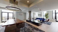 This single-floor space is designed with a contemporary aesthetic, offering a generously proportioned open-plan layout that seamlessly connects multiple functional zones. The main gathering area features light-toned hardwood flooring, recessed lighting, and large black-framed windows that fill the space with natural light and provide outward views. A bold cobalt blue sectional sofa anchors the central lounge area, complemented by mixed-material accent chairs, a round white coffee table, and gold side tables arranged atop a textured area rug. A prominent stone-clad structural column adds an architectural focal point.

Adjacent to the lounge, a dedicated dining area features a rich wood communal table with upholstered chairs beneath a striking circular pendant light. A textured accent wall displaying geometric hexagonal décor adds visual interest. The kitchenette nearby is outfitted with two-tone wood cabinetry, a mosaic tile backsplash, a sink, and integrated appliances.

A separate lounge nook features exposed white brick walls, framed artwork, a wall-mounted screen, and navy velvet armchairs arranged around a glass-and-brass coffee table — ideal for informal gatherings. Throughout, the space maintains a cohesive, sophisticated palette of navy, warm wood tones, and crisp whites.