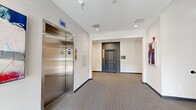 This well-appointed two-floor amenity space offers a thoughtfully designed environment for both arrival and recreation. The ground floor serves as the main entry point, featuring a bright, carpeted lobby with large glass double doors that invite natural light and views of the surrounding greenery. A carpeted staircase with sleek dark metal railings leads to the upper level, while a stainless steel elevator provides convenient accessibility between floors. The lower level also includes a carpeted corridor lined with framed artwork, accented doors, and functional utility space.

The upper floor expands into a generously proportioned recreation lounge with patterned carpet flooring and clean white walls. This social hub is equipped with a foosball table and a shuffleboard table, making it an ideal gathering space. A wall-mounted flat-screen TV adds a media component to the room, while multiple windows fitted with roller shades allow for adjustable natural light. Curated wall art and potted greenery contribute warmth and character throughout. Recessed lighting on both levels ensures a consistently bright and welcoming atmosphere, and the open staircase connection between floors enhances the sense of flow and vertical continuity throughout the space.