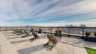 Located at 632 Newark Avenue in Jersey City, NJ, this expansive amenity floor offers an impressive blend of sophisticated interior spaces and dramatic outdoor terraces. The interior is defined by soaring floor-to-ceiling curtain wall windows that flood the space with natural light while framing sweeping views of the New York City skyline and surrounding waterfront. Light hardwood herringbone flooring runs throughout the open interior corridor, lending warmth and a refined aesthetic to the space.

The lounge area serves as a central gathering hub, featuring contemporary furnishings, large circular pendant light fixtures, and a wall-mounted display screen set against textured tile paneling. Adjacent seating nooks bathed in sunlight offer intimate settings for relaxation or collaboration, with direct visual access to the outdoor terrace. A dedicated office provides a private, functional workspace within the floor plan.

The rooftop terrace is a standout feature, offering generous open-air space finished with large-format porcelain pavers. A section of lush artificial turf is appointed with picnic tables and Adirondack-style seating, while a separate lounge zone features rows of modern reclining chaise lounges. The terrace is enclosed by sleek glass-panel railings, ensuring unobstructed panoramic views of the Manhattan skyline — making it an exceptional outdoor amenity in the heart of Jersey City.