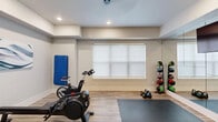 Located at 150 Country Squire Dr in Cromwell, Connecticut, this dedicated exercise room offers a thoughtfully designed fitness environment within a compact yet efficiently utilized space. The room features light wood-look flooring throughout, recessed ceiling lighting, and a neutral two-tone color scheme of soft gray and white that creates a clean, motivating atmosphere.

A full wall of floor-to-ceiling mirrors amplifies the sense of space and provides visual feedback during workouts. A wall-mounted flat-screen television adds an entertainment option for those looking to stay engaged during longer sessions. The room is equipped with a diverse range of fitness equipment, including cycling bikes, a rowing machine, kettlebells, medicine balls on a dedicated rack, resistance bands, TRX suspension trainers, and balance tools such as a stability ball and step platforms.

Exercise mats are laid out to define a stretching and floor workout zone, while a blue gymnastics mat mounted on the wall offers additional versatility. A roller window shade allows natural light to be adjusted as needed. The space is accessed through stylish double glass-panel doors in a navy frame, contributing to its polished, professional aesthetic. Decorative artwork on the walls adds a finished touch, making this fitness space as visually appealing as it is functional.