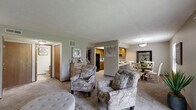 Located at 4880 Willow Glen Dr in Beech Grove, Indiana, this single-story, 3-bedroom, 2-bathroom home offers a practical and comfortable layout across its thoughtfully designed floor plan. The living room serves as the heart of the home, featuring plush carpeting, a textured ceiling, and a sliding glass door that invites natural light and provides outdoor access. Adjacent to the living room, the dedicated dining area accommodates a round table and chairs, with a window overlooking the outdoors and warm neutral-toned walls throughout. The kitchen is well-appointed with warm wood cabinetry, granite-style countertops, black appliances including a refrigerator, electric range, microwave, and dishwasher, plus light vinyl flooring and a skylight-style ceiling detail that brightens the space. The primary bedroom offers generous proportions, soft carpet, and a large built-in closet with wood panel doors. Additional bedrooms are similarly carpeted and bright, with windows providing natural light. The two bathrooms include a full bath with a tub-shower combination and wood-toned vanity cabinetry, and a second bath with a wide vanity and mirror. Throughout the home, wood trim accents and neutral gray walls create a cohesive, welcoming ambiance.