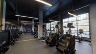 Located at 160–172 Fourth St in Oakland, CA, this single-floor fitness center offers a comprehensive workout environment designed with both functionality and aesthetic in mind. The space features an industrial-inspired design, characterized by exposed concrete columns, dark painted ceilings with visible structural elements, and sleek linear pendant lighting that runs throughout the facility, creating a bold, high-energy atmosphere.

The cardio zone is positioned along the perimeter, taking advantage of large floor-to-ceiling windows that allow ample natural light and provide street-level views of the surrounding Oakland neighborhood. Treadmills, stair climbers, ellipticals, and stationary bikes are arranged in organized rows, offering a well-stocked selection for cardiovascular training.

A dedicated strength training area features cable machines, multi-use racks, and a dumbbell section lined with full-length mirrors accented by yellow trim details. Rubber flooring in this zone provides durability and safety. A colorful, large-scale athletic mural adds a dynamic visual element to one wall, reinforcing the facility's energetic character.

The overall design incorporates a consistent dark color palette with yellow accent details, giving the space a cohesive, contemporary identity suited to a modern urban fitness environment.